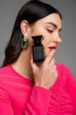 portrait of alluring young woman with brunette hair and red lips posing in trendy magenta dress while holding bottle of luxurious perfume isolated on grey background  clipart