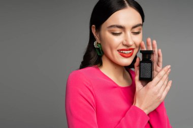 charming young woman with shiny brunette hair, red lips posing in magenta dress while holding bottle of perfume and smiling isolated on grey background  clipart