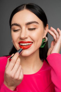 Moda küpeleri ve esmer saçları olan, kırmızı ruj süren, yüzünü çeviren ve gri arka planda yalnız poz veren muhteşem bir kadının portresi.