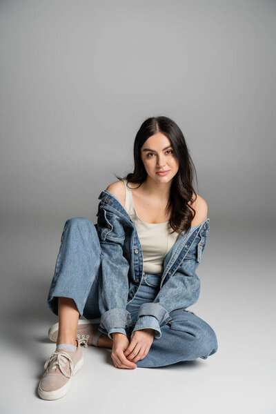 full length of young charming woman with flawless natural makeup posing in stylish blue jeans and denim jacket while sitting and looking at camera on grey background