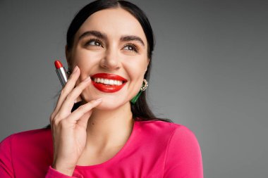 Moda küpeleri olan güzel bir kadının portresi. Parlak esmer saçlı, parmaklarının arasında kırmızı ruj tutan ve gri arka planda poz verirken gülümseyen bir kadın. 