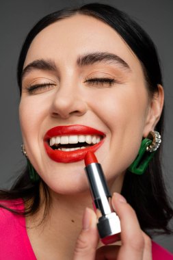 Moda küpeleri, parlak esmer saçları olan, lüks kırmızı ruj süren ve gri arka planda poz verirken gülümseyen şık genç bir kadının yakın görüntüsü. 