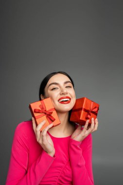 alluring young woman with shiny brunette hair smiling while standing in magenta party dress and holding wrapped gift boxes for holiday on grey background  clipart