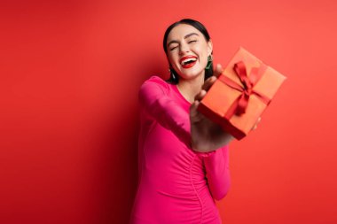 Heyecanlı, esmer ve moda küpeli bir kadın. Mavi parti elbisesi içinde dikilirken gülümsüyor ve kırmızı arka planda tatil için paketlenmiş hediye kutusunu tutuyor. 