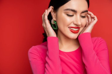 joyful and shy woman in trendy earrings adjusting her brunette hair and smiling while standing in magenta party dress while posing on red background clipart