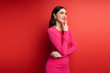 Saçları esmer ve son moda küpeli güzel bir kadın. Kırmızı arka planda durup poz verirken ve bakarken gülümsüyor.