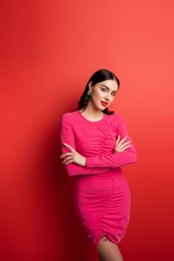 gorgeous woman with brunette hair and trendy earrings standing with folded arms in magenta party dress while posing and looking at camera on red background clipart