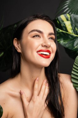 Kumral ve kırmızı dudakları olan mutlu genç bir kadın, elleri göğsünde, ıslak ve yeşil palmiye yapraklarının etrafında, yağmur damlalarıyla gülümserken ve başka tarafa bakarken gülümsüyor.