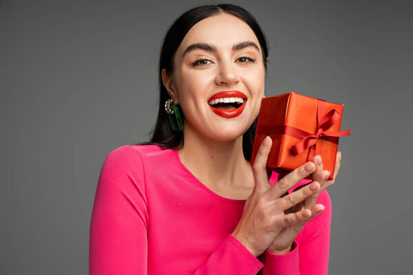 возбужденная и шикарная молодая женщина с модными серёжками и безупречным макияжем, улыбающаяся, держа красную и обернутую подарочную коробку на сером фоне 