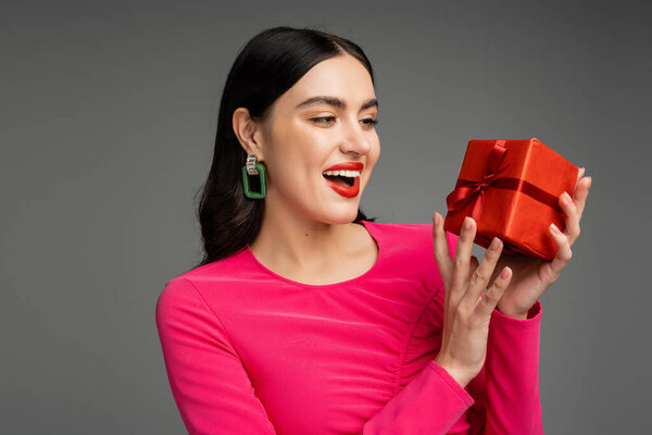 возбужденная и шикарная молодая женщина с модными серёжками и блестящими волосами брюнетки, улыбающаяся, держа на сером фоне красную и обернутую подарочную коробку 