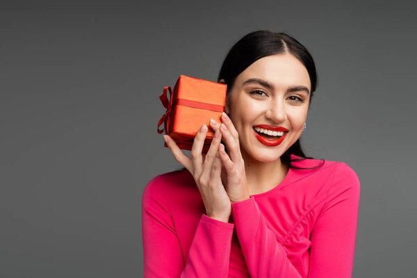 позитивная и шикарная молодая женщина с модными серёжками и блестящими брюнетками, улыбающаяся, держа на сером фоне красную и обернутую подарочную коробку 
