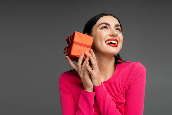 счастливая молодая женщина с модными серёжками и блестящими волосами брюнетки улыбается, держа в руках красную и обернутую подарочную коробку на отдыхе и глядя вверх на сером фоне 