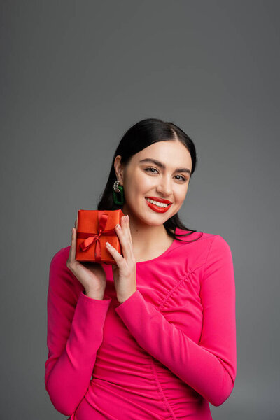 элегантная и молодая женщина с модными сережками и брюнетками улыбается, держа красный и обернутый подарок для отдыха на сером фоне 
