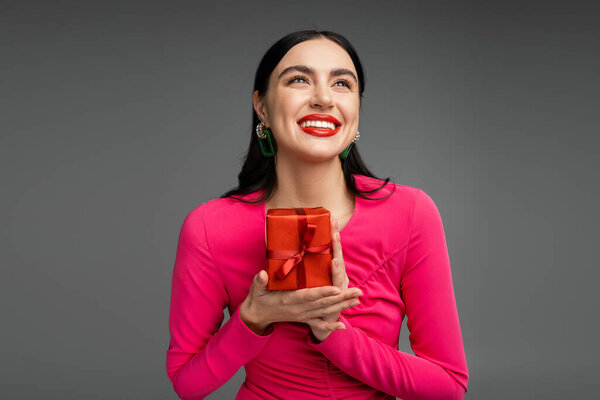 elegant and young woman with brunette hair and trendy earrings smiling while looking up and holding red and wrapped present for holiday on grey background 