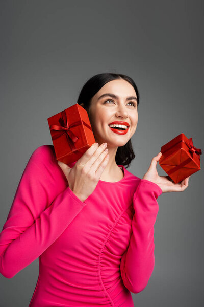 веселая молодая женщина с брюнетками и модными серёжками, улыбающаяся, стоя в пурпурном платье для вечеринок и держа упакованные подарочные коробки для отдыха на сером фоне 