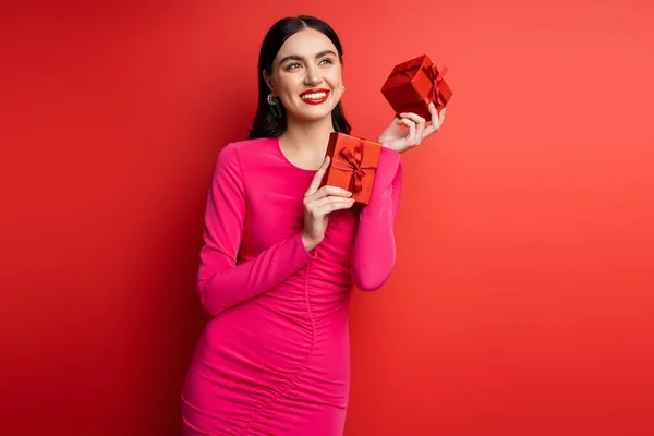穿着洋红色派对礼服 拿着包装好的礼品盒在红色背景下度假的迷人女人 一头乌黑的头发 戴着时髦的耳环 面带微笑 — 图库照片