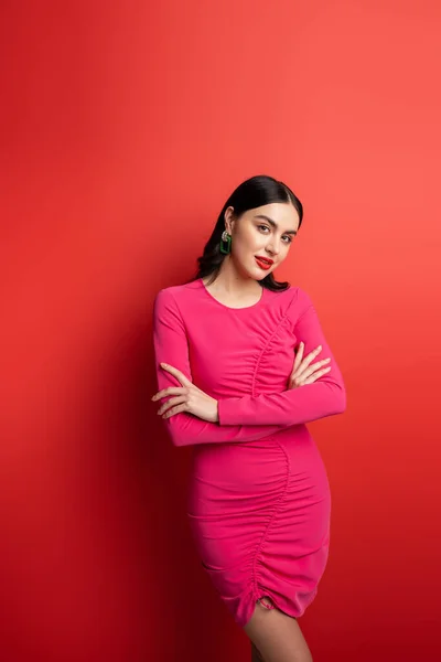 stock image gorgeous woman with brunette hair and trendy earrings standing with folded arms in magenta party dress while posing and looking at camera on red background