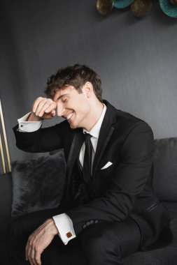 Beyaz gömlekli ve kravatlı mutlu ve genç damat portresi. Yüzüne dokunurken gülümsüyor ve modern otel odasındaki rahat, gri koltukta düğün günü oturuyor.