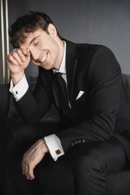 portrait of positive young groom in black suit with white shirt and tie touching face with hand while smiling and sitting on comfortable dark grey couch in modern hotel room on wedding day clipart