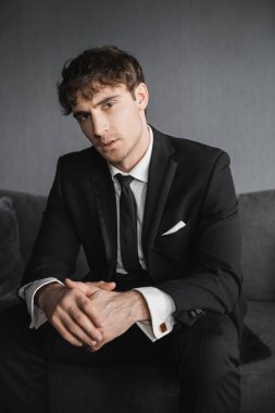 Beyaz gömlekli ve kravatlı yakışıklı genç damat portresi. Koyu gri koltukta oturuyor ve düğün günü modern otel odasında kameraya bakıyor.