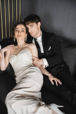 charming young bride in luxurious earrings with pearls and white wedding dress and good looking groom in black suit with tie sitting together on dark grey couch in hotel room and looking at camera  clipart