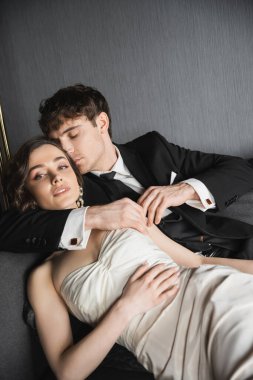 good looking groom in black suit with tie hugging and kissing bride in earrings with pearls and necklace lying in white wedding dress on dark grey couch in hotel room clipart