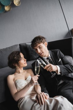 happy newlyweds, bride in white wedding dress and groom in black suit holding glasses of champagne while clinking and celebrating their marriage after wedding in hotel room   clipart