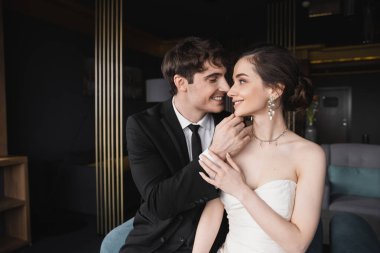 Modern otel süitinde birbirlerine bakarken beyaz gelinlik ve mücevherlerle etkileyici bir yüze sahip siyah takım elbiseli mutlu bir damat.