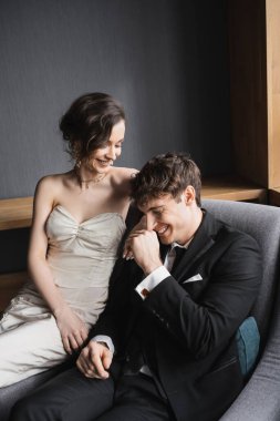Mutlu gelin, beyaz gelinlik ve lüks takılar içinde gülümsüyor ve siyah takım elbiseli pozitif damat ile birlikte otel odasındaki rahat koltukta oturuyor. 