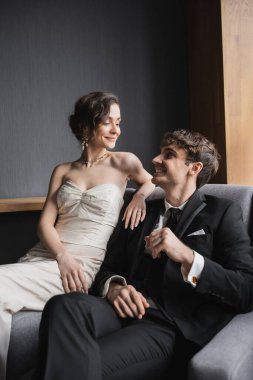 Beyaz gelinlik içinde mutlu bir gelin ve lüks bir kolye içinde gülümserken siyah takım elbiseli pozitif bir damat otel odasında rahat bir koltukta oturuyor. 