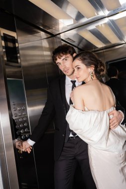 good looking groom in formal wear pushing button while hugging brunette bride in wedding dress and standing together in elevator of modern hotel  clipart