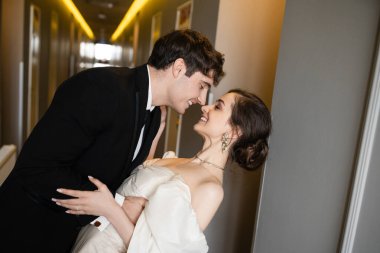 side view of happy groom in suit leaning towards charming bride in white wedding dress while smiling together in corridor of modern hotel, couple on honeymoon   clipart