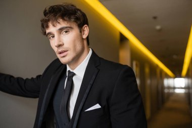 Düğünde modern otelin koridorunda dikilirken gömlek ve kravat takan siyah elbiseli yakışıklı ve genç damat portresi. 