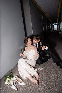 Siyah takım elbiseli mutlu bir damat elinde şişe, elinde bir bardak şampanyayla muhteşem bir gelinin yanında oturuyor ve modern otelin koridorunda topuklu ayakkabılar, balayında yeni evliler. 