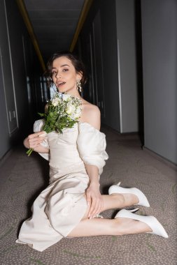 amazed young bride with brunette hair in white wedding dress looking at camera while holding bridal bouquet with flowers and sitting on floor of hallway in modern hotel  clipart