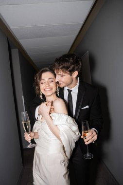 Beyaz gelinlik içinde genç ve neşeli bir gelin ve elinde şampanya bardakları olan neşeli bir damat otelin koridorunda birlikte durup gülümserken, yeni evliler balayında. 