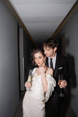 Beyaz gelinlik içinde genç ve esmer gelin ve elinde şampanya bardakları olan yakışıklı damat otelin koridorunda birlikte duruyorlar, yeni evliler balayında. 