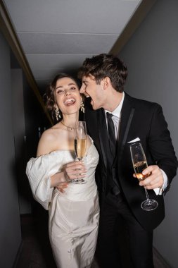 excited groom hugging young and brunette bride in white wedding dress and holding glasses of champagne while standing and smiling together in hallway of hotel, newlyweds on honeymoon  clipart