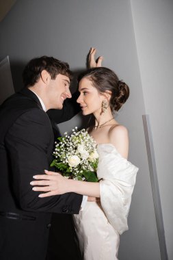 happy groom in black suit leaning towards wall and looking at bride in white wedding dress holding bridal bouquet while standing together in hallway of modern hotel  clipart