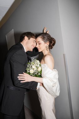 positive groom in black suit leaning towards wall near bride in white wedding dress holding bridal bouquet while standing together in hallway of modern hotel, newlyweds on honeymoon  clipart