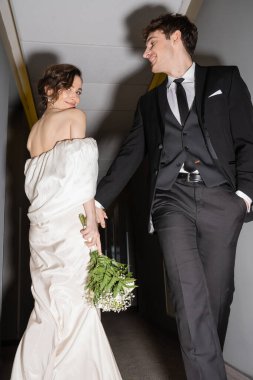 low angle view of cheerful groom in black suit posing with hand in pocket and looking at happy bride in white wedding dress holding bridal bouquet while walking together in hall of modern hotel  clipart