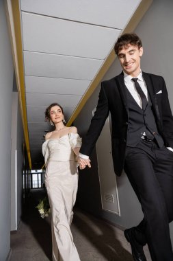 low angle view of positive groom in black suit posing and holding hands with bride in white dress carrying bridal bouquet while walking together in corridor of modern hotel  clipart