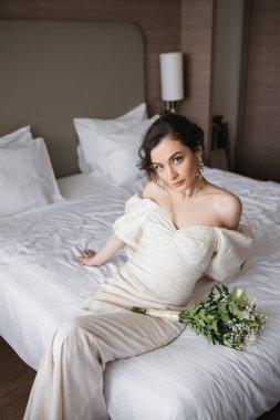 Beyaz elbiseli genç gelin ve lüks takılar. Çiçekli gelin buketinin yanında oturuyor ve düğün günü otel odasındaki modern yatak odasında kameraya bakıyor. 