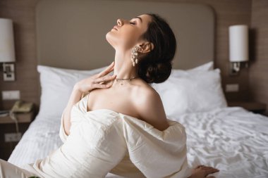Beyaz elbiseli, inci küpeli, kolyeli ve lüks takılı gelinin yan görünüşü. Düğün günü modern bir yatak odasında yatağa yaslanırken. 