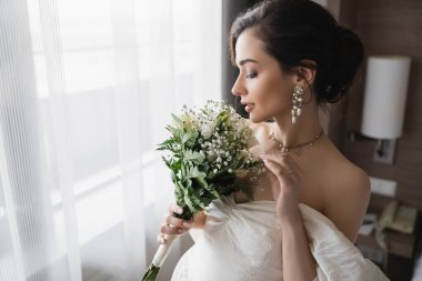 side view of elegant young bride in white dress and luxurious jewelry with pearl earrings holding bridal bouquet in modern hotel room on wedding day  clipart