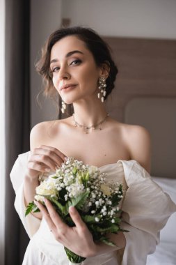 Büyüleyici gelinlik ve lüks takılar takan, çiçekli gelin buketi tutan ve düğün günü otel odasındaki modern yatak odasındaki kameraya bakan büyüleyici genç kadın. 