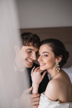 Mücevher ve gelinlik içinde güzel genç bir gelin klasik resmi giyinmiş neşeli damadın omzuna sarılıyor ve modern otel odasında seremoni üstüne tören yapıyor. 