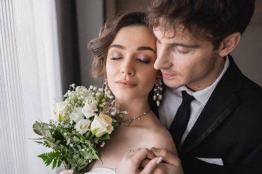 Mücevher içinde güzel bir gelin, geleneksel resmi kıyafetlerle damat ile el ele tutuşan beyaz bir elbise. Düğün töreninden sonra modern otel odasında kapalı gözlerle yan yana dururken. 