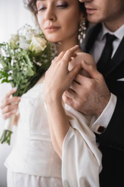 Mücevher içindeki genç gelin, gelin buketi olan beyaz elbise, geleneksel resmi kıyafetlerle damat ile el ele tutuşurken düğün töreninden sonra modern otel odasında yan yana duruyoruz. 