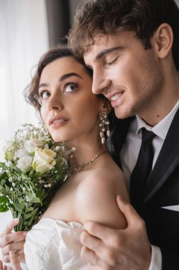 happy groom in classic formal wear touching hand of pretty young bride in jewelry, white dress with bridal bouquet while standing together in modern hotel room after wedding ceremony  clipart